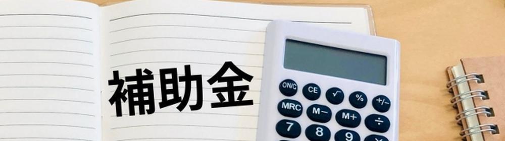 木目のテーブルの上に置かれたノートに「補助金」と書かれており、隣には電卓と白いペンが添えられた、家計や経済支援をイメージさせる写真。画像全体は白い余白に黒枠が加えられている。