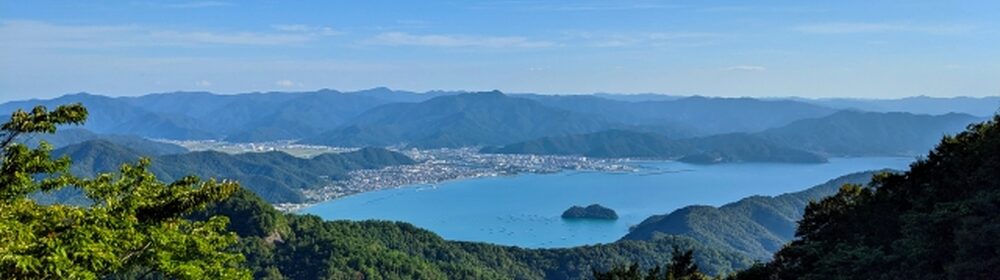 緑豊かな山の斜面越しに、静かな湖と湖上の小島、遠くに連なる山々と青空が広がる風景写真