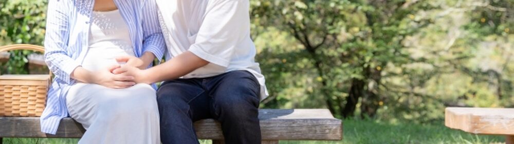 ベンチに座り、妊婦の女性の膝に手を添える男性の上半身が映る、公園の木陰で穏やかに寄り添うカップルの写真