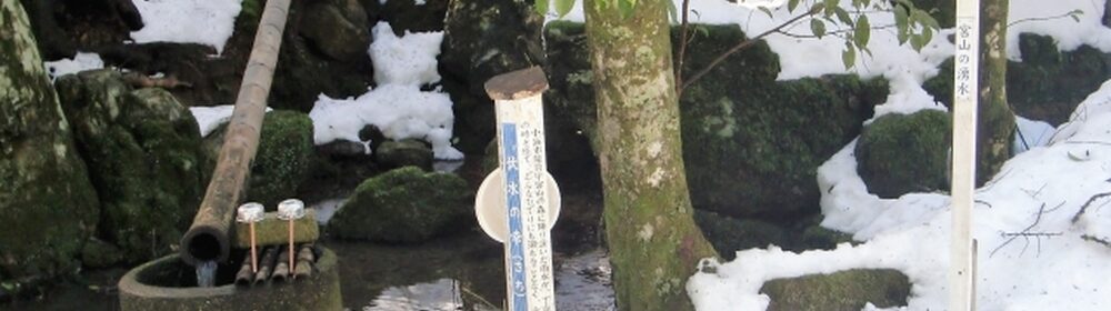雪に覆われた地面の上に石製の手水鉢と柄杓、手水と書かれた案内板が立つ、冬の神社の手水場の風景