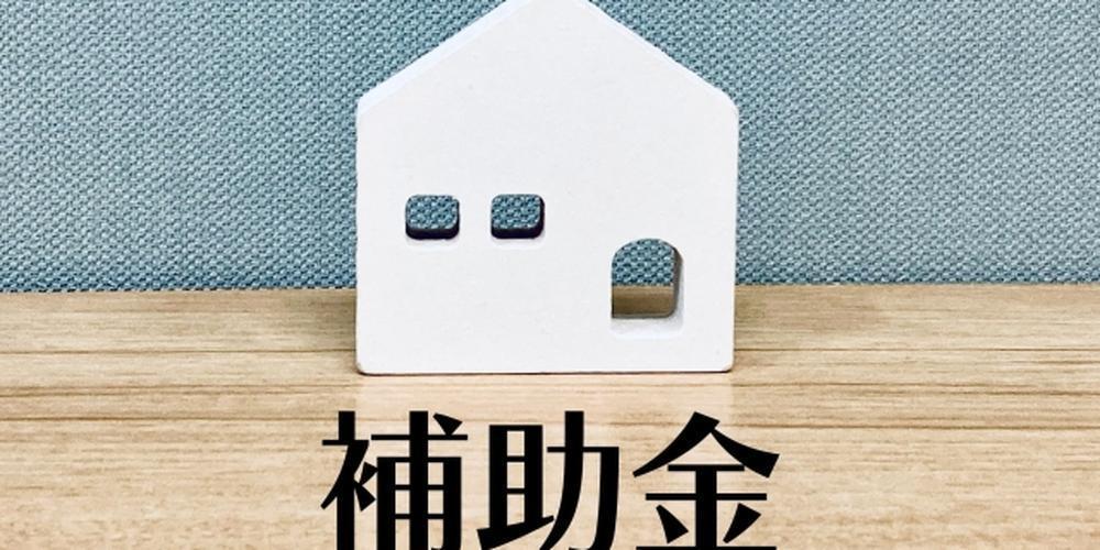 木目のテーブルの上に置かれた白い家型のオブジェと、その下に大きく「補助金」と書かれた文字。背景は青系のファブリック風で、画像の外側は黒い枠で囲まれている。